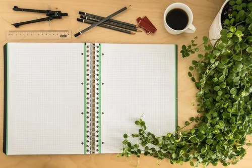 Un carnet de notes ouvert et posé sur un bureau. Il montre des pages vierges. Des crayons de bois, un compas, un café et une plante verte sont posés à côté - apprendre à gérer ses inquiétudes - Hypnose - Coaching de vie
