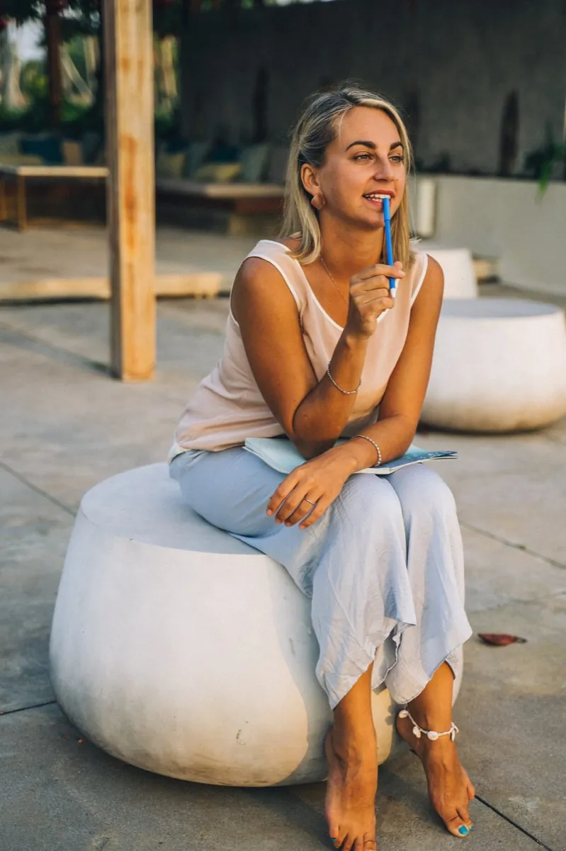 Une femme, pieds nus, assise à l'extérieur, réfléchit avec un stylo à la main et un carnet ouvert sur les genoux - inquiétudes - stress - anxiété - hypnose