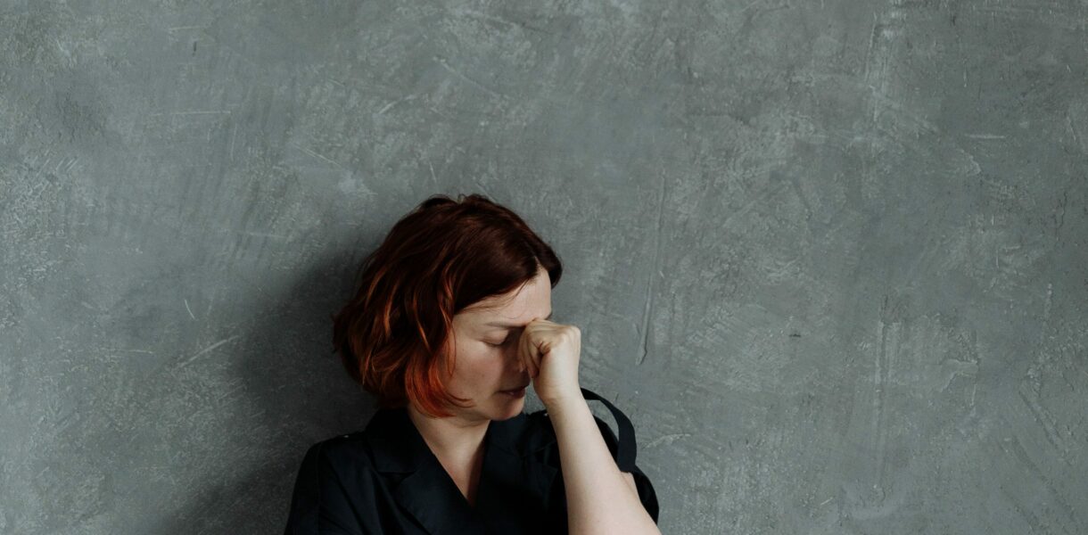 Une femme seule contre un mur de béton gris se tient le visage, entre les deux yeux. Le coaching et l'hypnose peuvent l'aider à gérer ses inquiétudes.