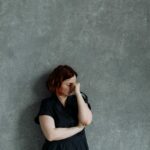 Une femme seule contre un mur de béton gris se tient le visage, entre les deux yeux. Le coaching et l'hypnose peuvent l'aider à gérer ses inquiétudes.