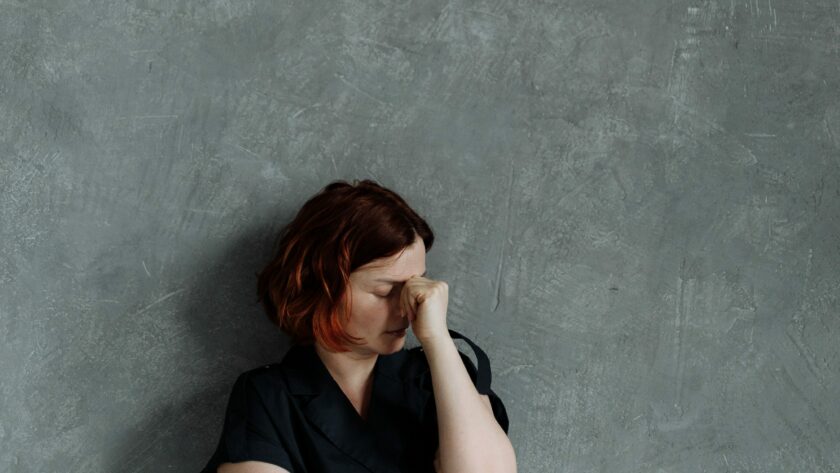 Une femme seule contre un mur de béton gris se tient le visage, entre les deux yeux. Le coaching et l'hypnose peuvent l'aider à gérer ses inquiétudes.