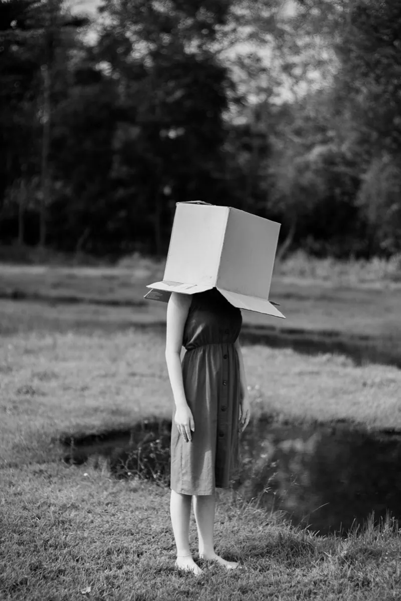 Photo en noir et blanc d'une jeune femme debout dans un parc avec un carton qui cache sa tête - syndrome de l'imposteur - coaching professionnel