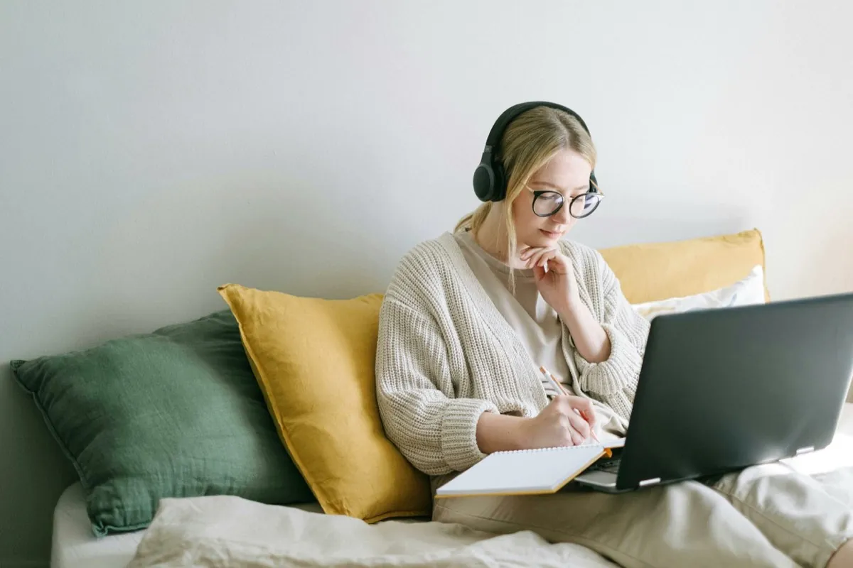 Une jeune femme assise sur son lit travaille avec un casque sur les oreilles, un ordinateur sur les genoux et un stylo à la main - coaching de vie - hypnose à Nantes - Sylvie Rauwel - Nantes
